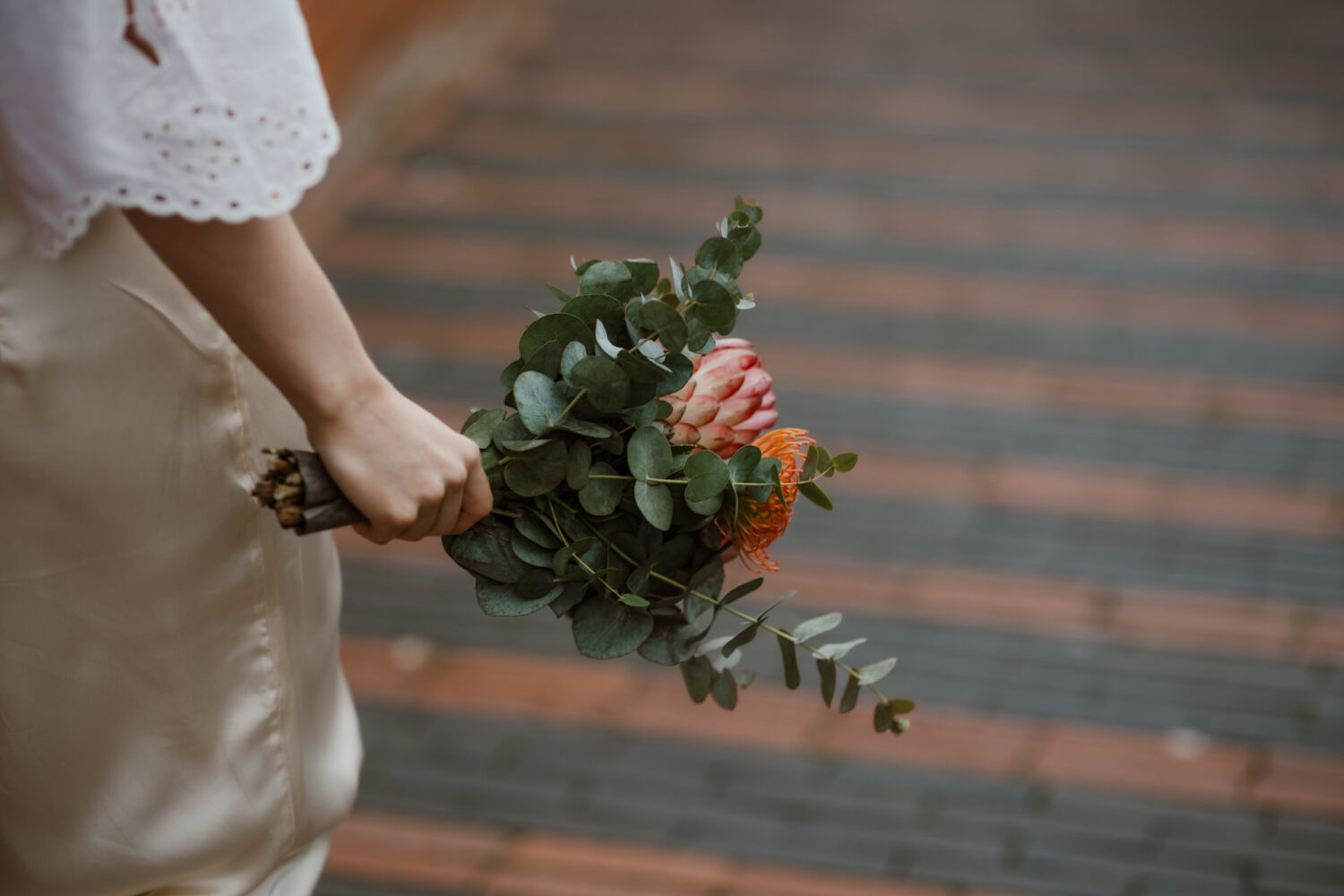 Person holding a bouquet with eucalyptus leaves and a pink protea flower, standing on a brick path.