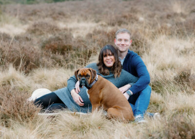 A couple sitting on grass in a field, with a brown dog in front of them. They are outdoors, surrounded by dry, beige plants.