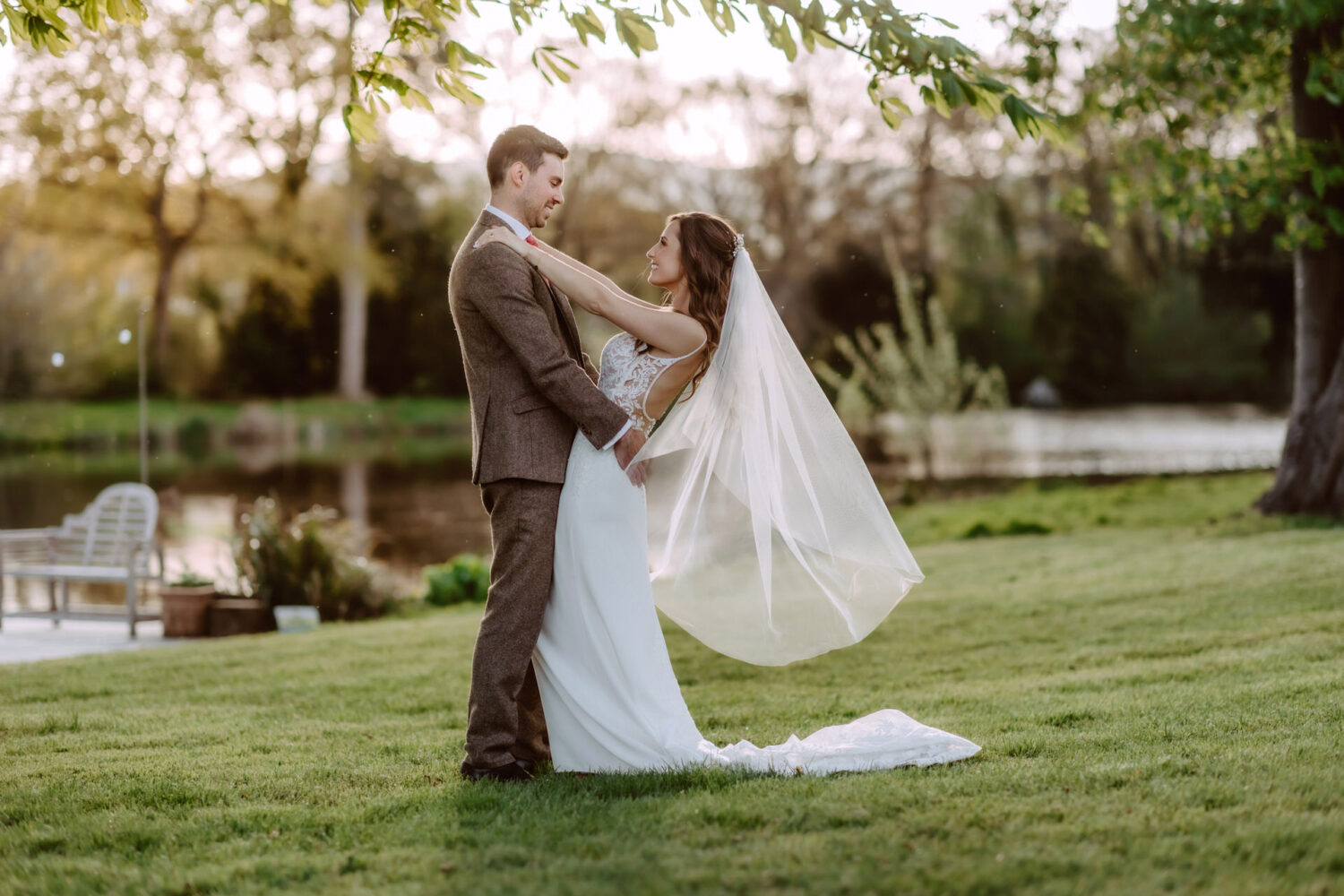 A bride and groom stand on grass near a pond, embracing each other. The bride's veil flows behind her. Trees are in the background.