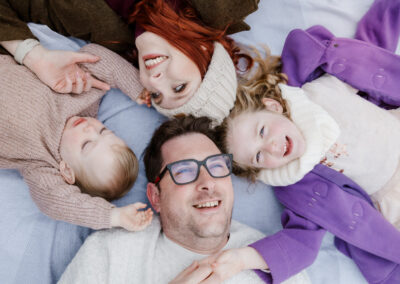 A family of four, wearing cozy clothing, lies on a blanket outdoors. They are smiling and looking up at the camera, with two children positioned between two adults.