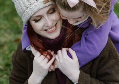 Woman wearing a beanie and coat, sitting on grass, with a child in a purple jacket hugging her from behind.