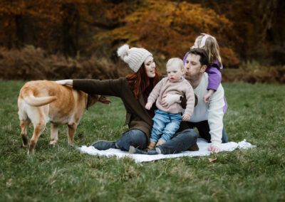 Family sitting on grass with a blanket; woman holding a baby, man kissing toddler. Dog standing nearby.