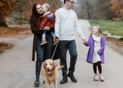 Family walking on a forest path with a dog. A woman holds a toddler, a man holds a child's hand, and the dog walks in front. The path is surrounded by autumn trees.