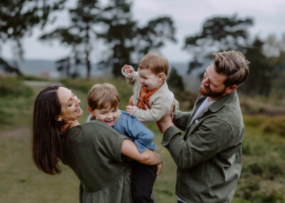 A family of four laughing outdoors. A woman holds a child, while a man lifts another child. Trees and a grassy field are in the background.