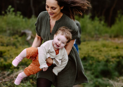 Woman in a green dress carries a smiling toddler in an orange outfit, surrounded by greenery.