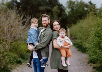 A family of four stands on a gravel path surrounded by trees. The father holds a young boy, while the mother carries a baby. Both adults smile at the camera.