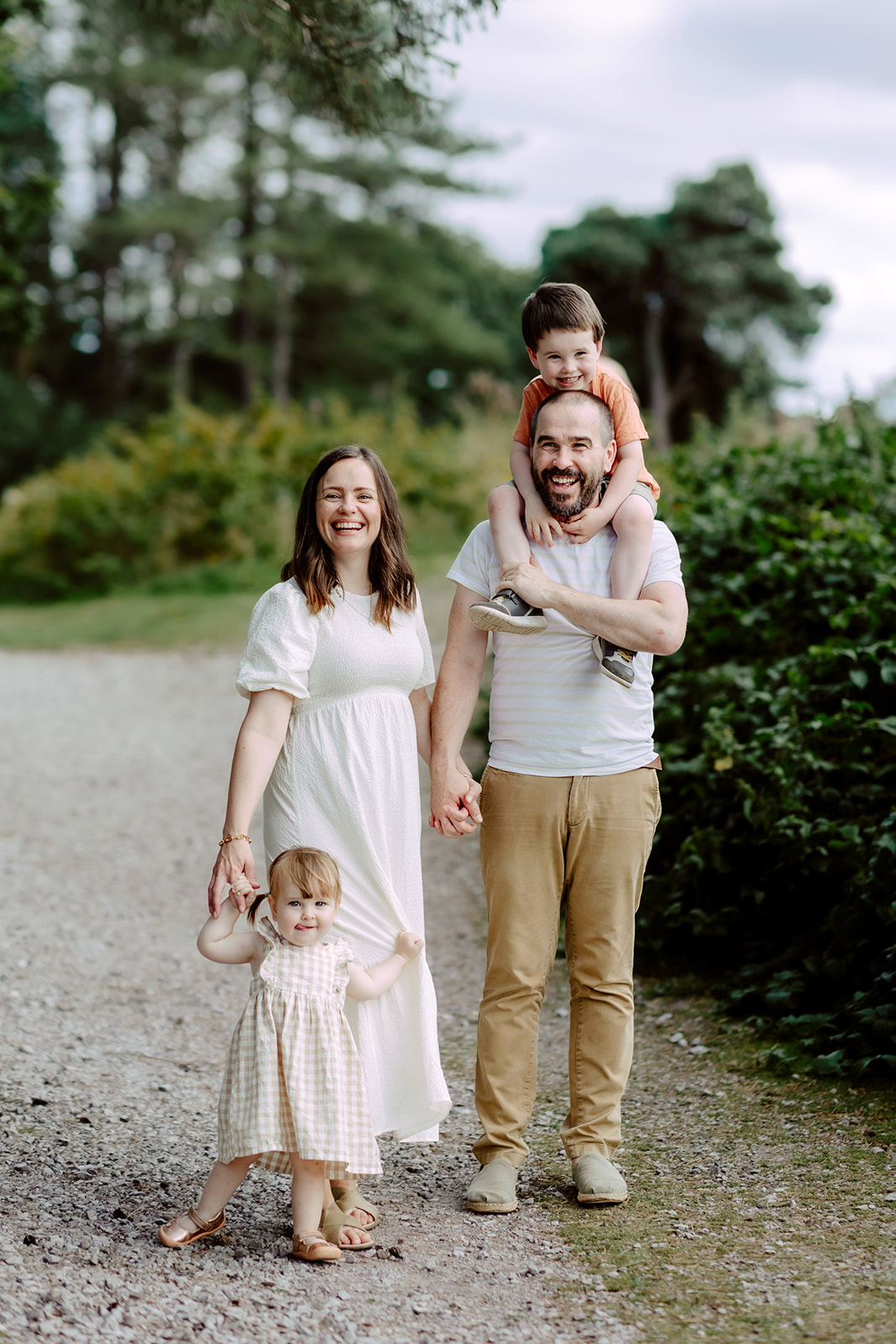 A family of four stands on a gravel path in a park. The father holds a child on his shoulders. The mother holds hands with a toddler. They are all smiling. Trees and bushes surround them.