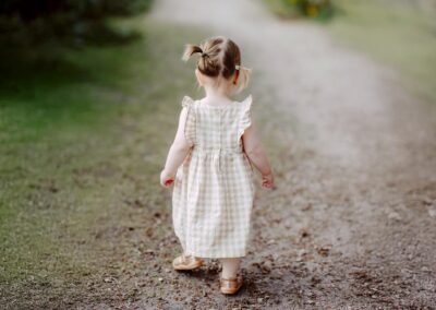 A toddler with pigtails in a plaid dress walks away on a dirt path surrounded by grass.