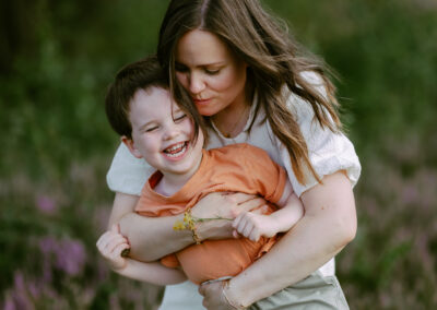 A woman hugs a laughing child from behind in a field, both smiling. The child wears an orange shirt and holds yellow flowers.