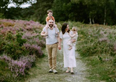 A family of four walks along a path. The father carries a boy on his shoulders, the mother holds a girl's hand. Green and purple foliage surrounds them.