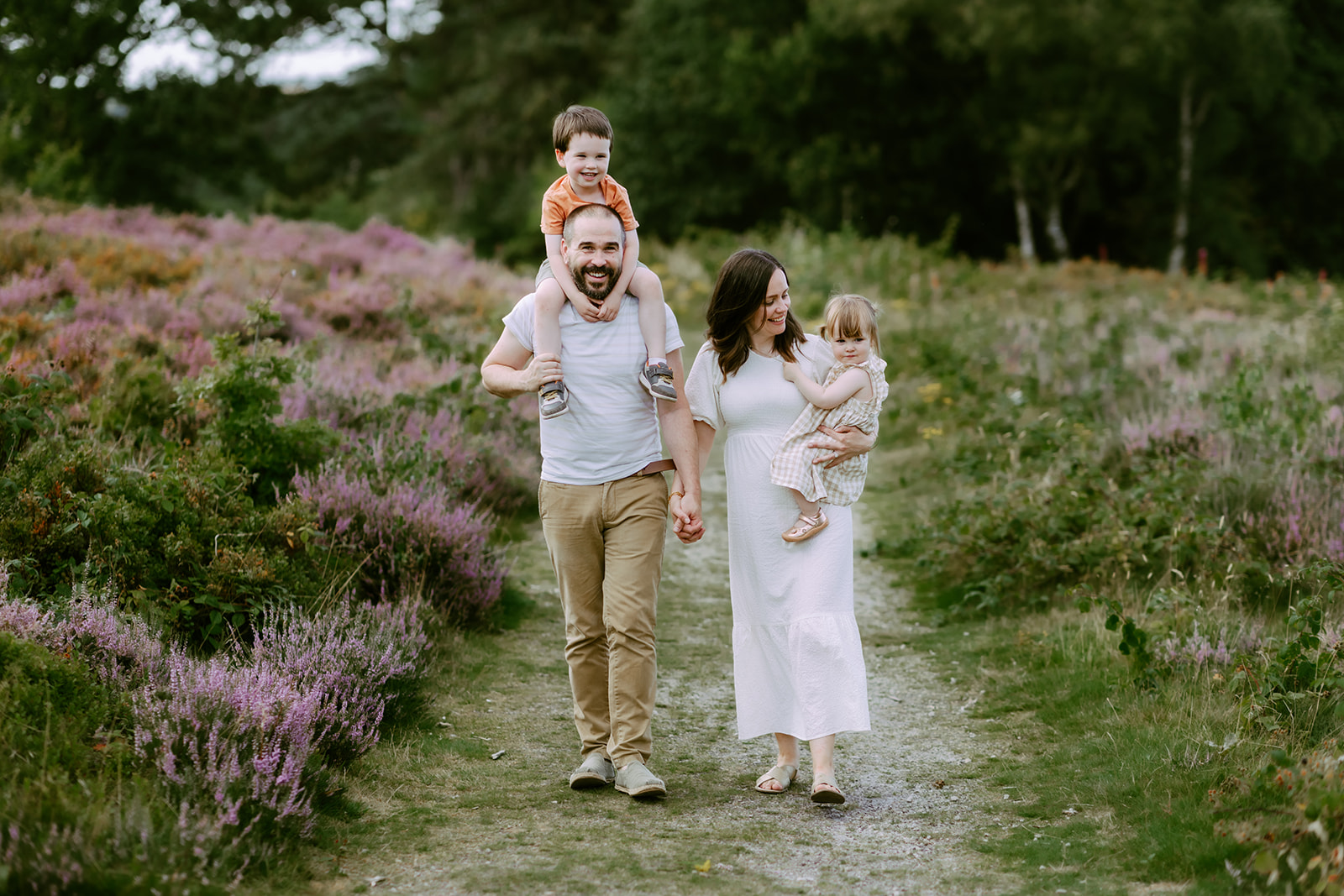 A family of four walks along a path. The father carries a boy on his shoulders, the mother holds a girl's hand. Green and purple foliage surrounds them.