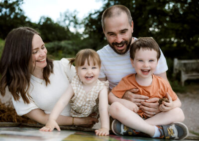 A family of four, two adults and two young children, smile outdoors. The adults hold the children, who are dressed casually. Trees and a path are in the background.