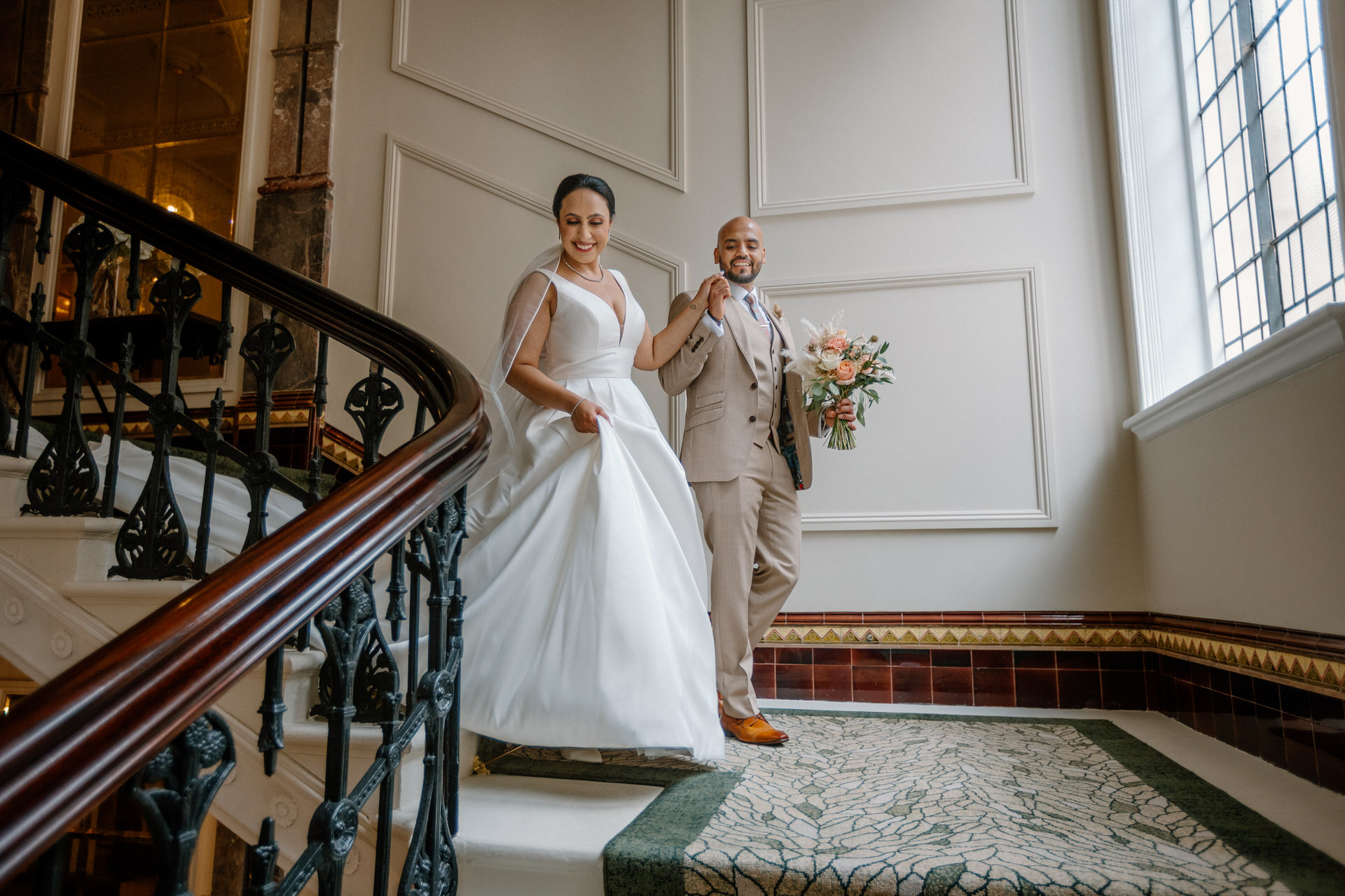 A bride in a white dress and a groom in a tan suit walk down a staircase in The Grand Hotel Birmingham, holding hands and smiling, beautifully captured by their wedding photographer.