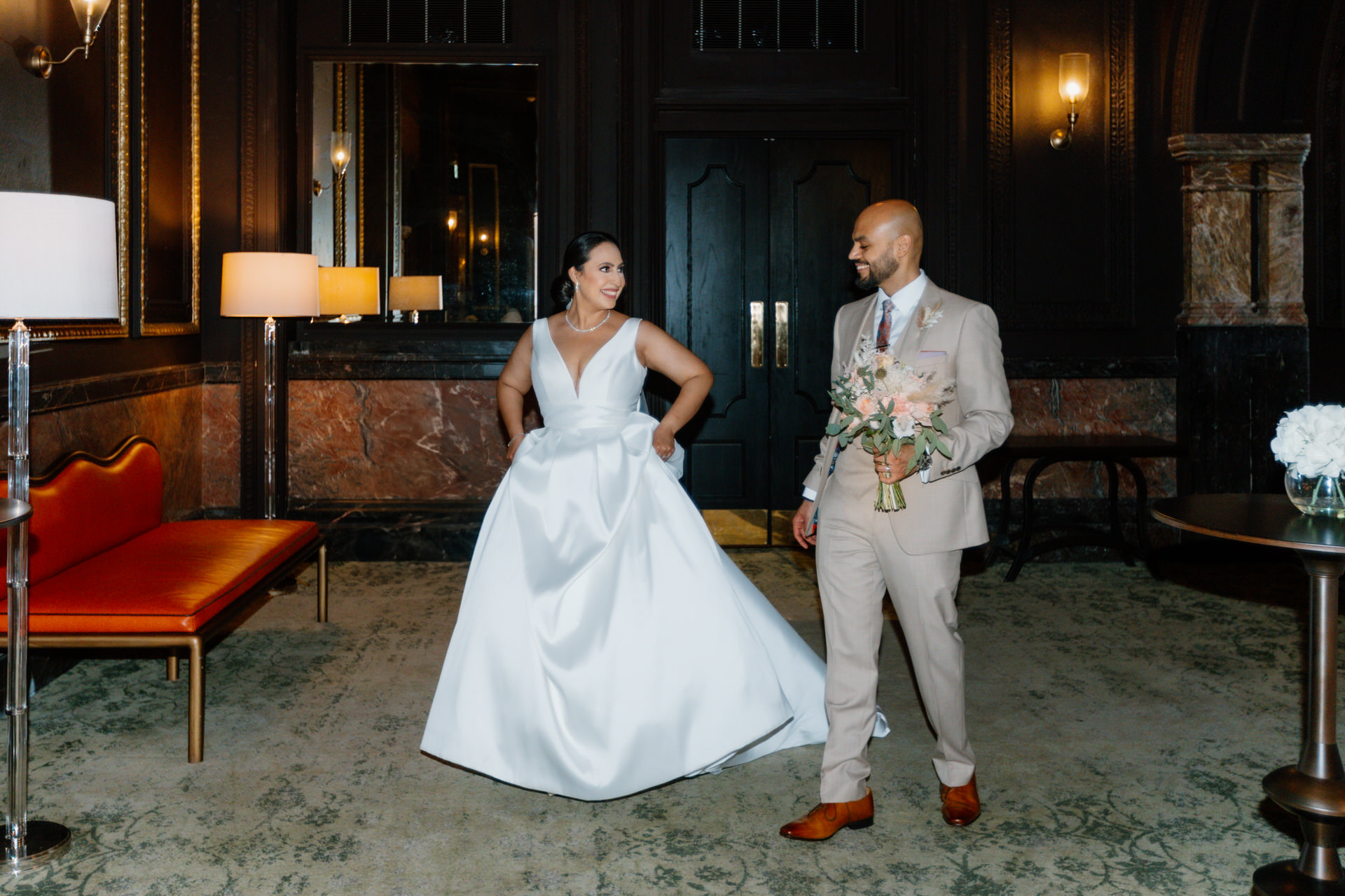 A bride in a white gown and a groom in a tan suit holding a bouquet stand together in an elegant, dimly lit room with dark walls and marble accents at The Grand Hotel Birmingham.