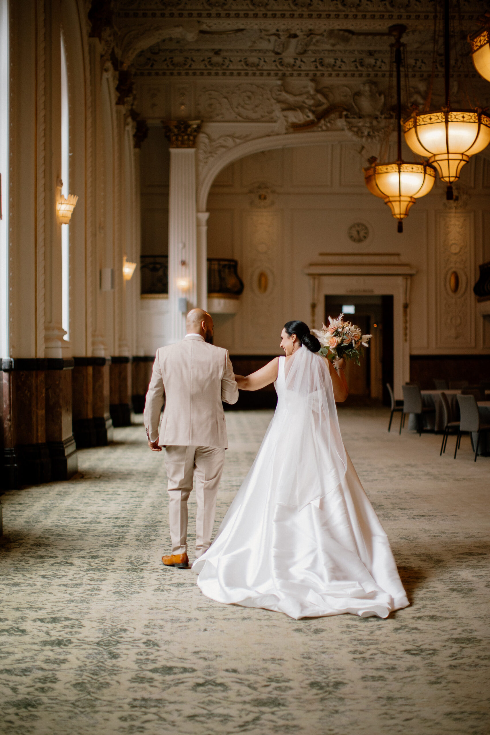 A bride and groom walk together in an elegant, high-ceilinged room with large windows and ornate chandeliers at The Grand Hotel Birmingham.