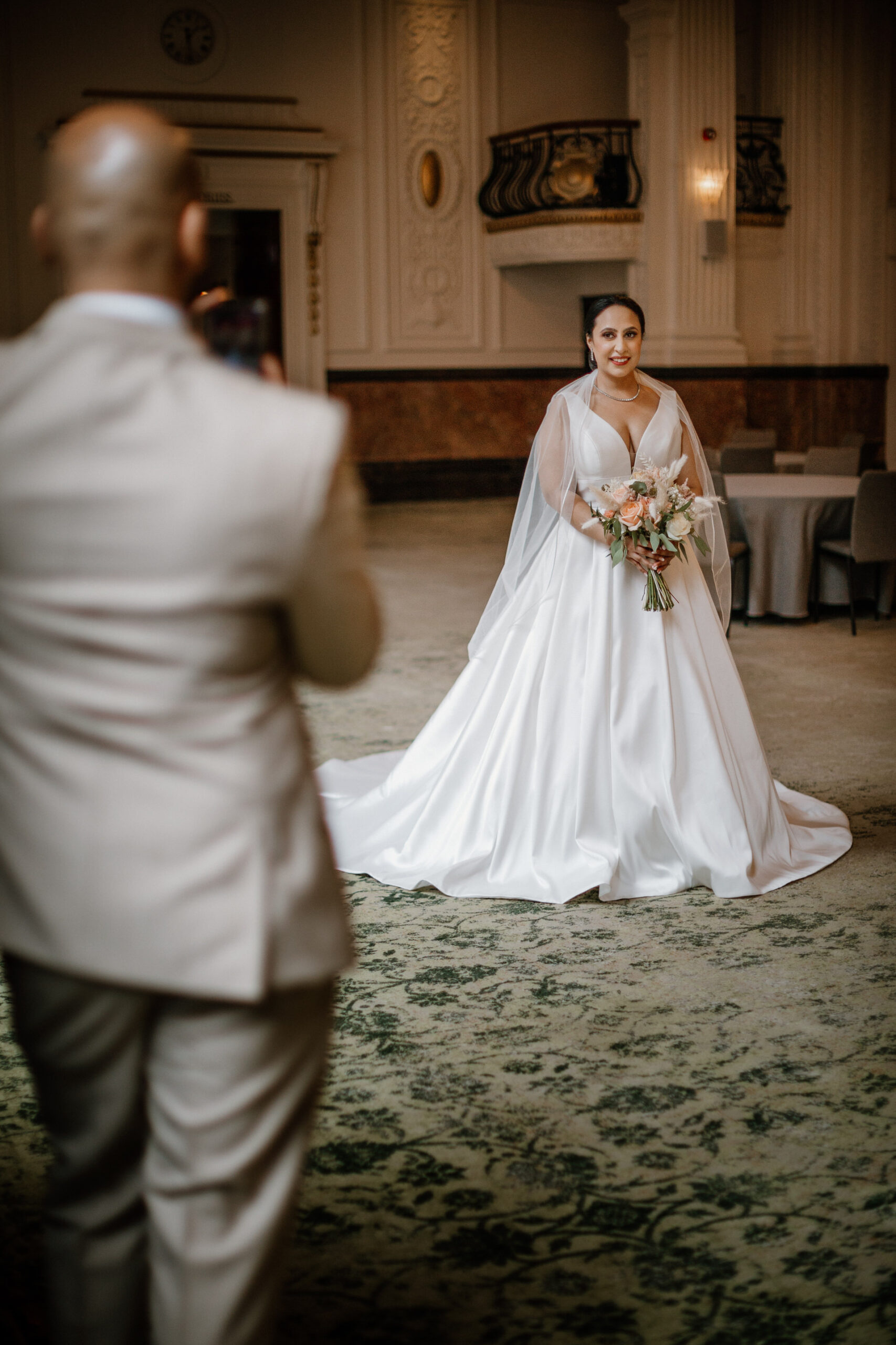 A bride in a white gown holding a bouquet stands indoors at a top venue in Birmingham, while a man in a light suit faces her, appearing to take a photo.