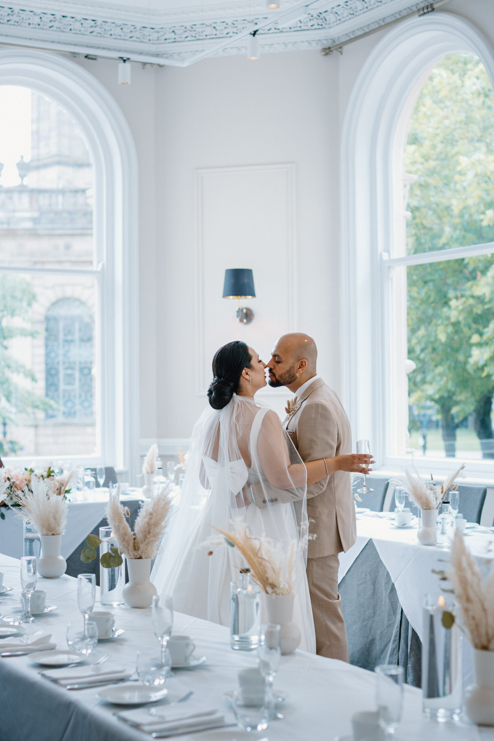 A bride and groom share a kiss while standing at the head of a decorated table in a bright, elegant room with large windows and natural light at their grand hotel wedding.