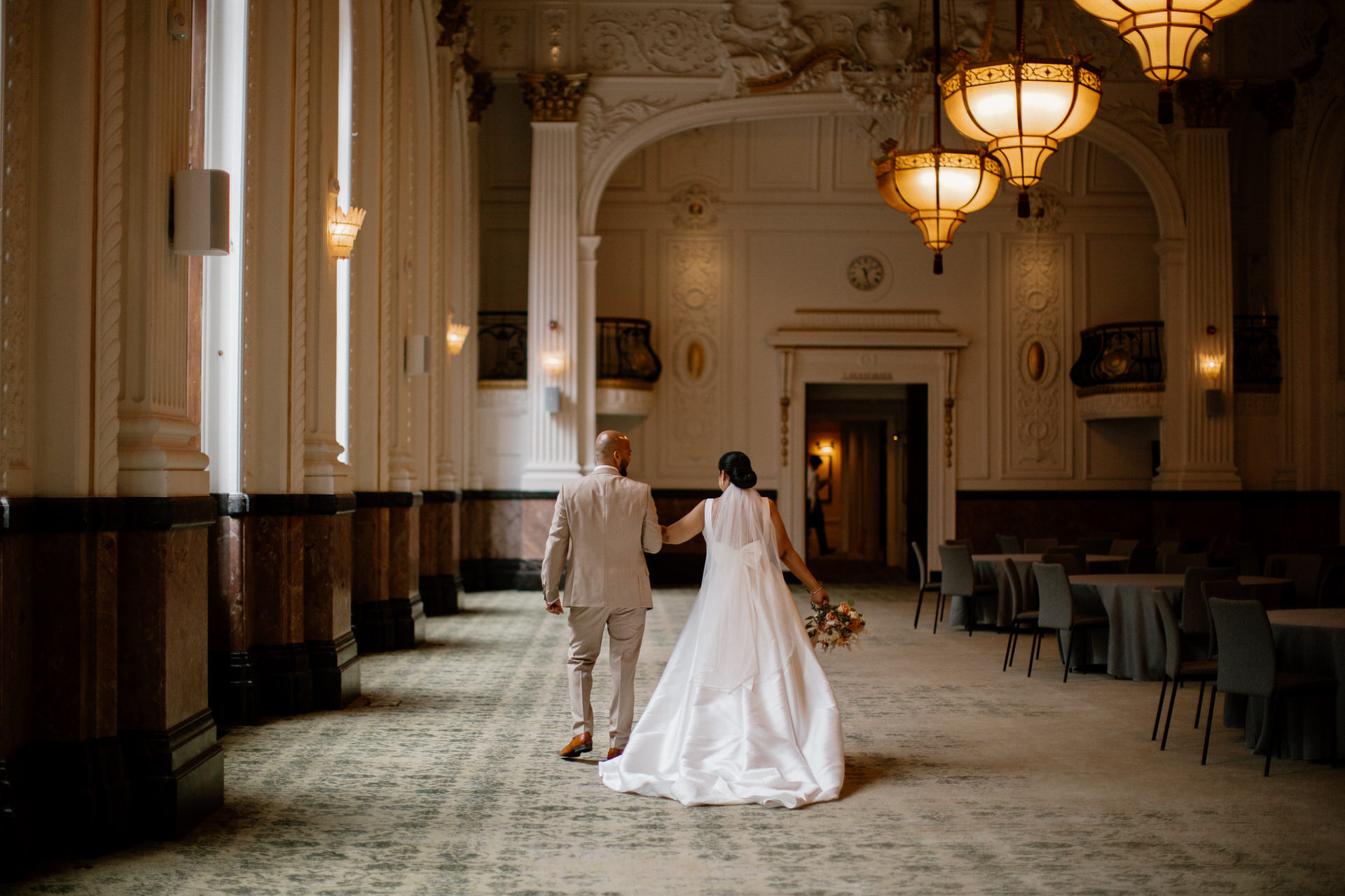 A bride and groom walk arm in arm through the elegant, chandelier-lit hall of The grand Hotel Birmingham, surrounded by high ceilings and ornate decor.