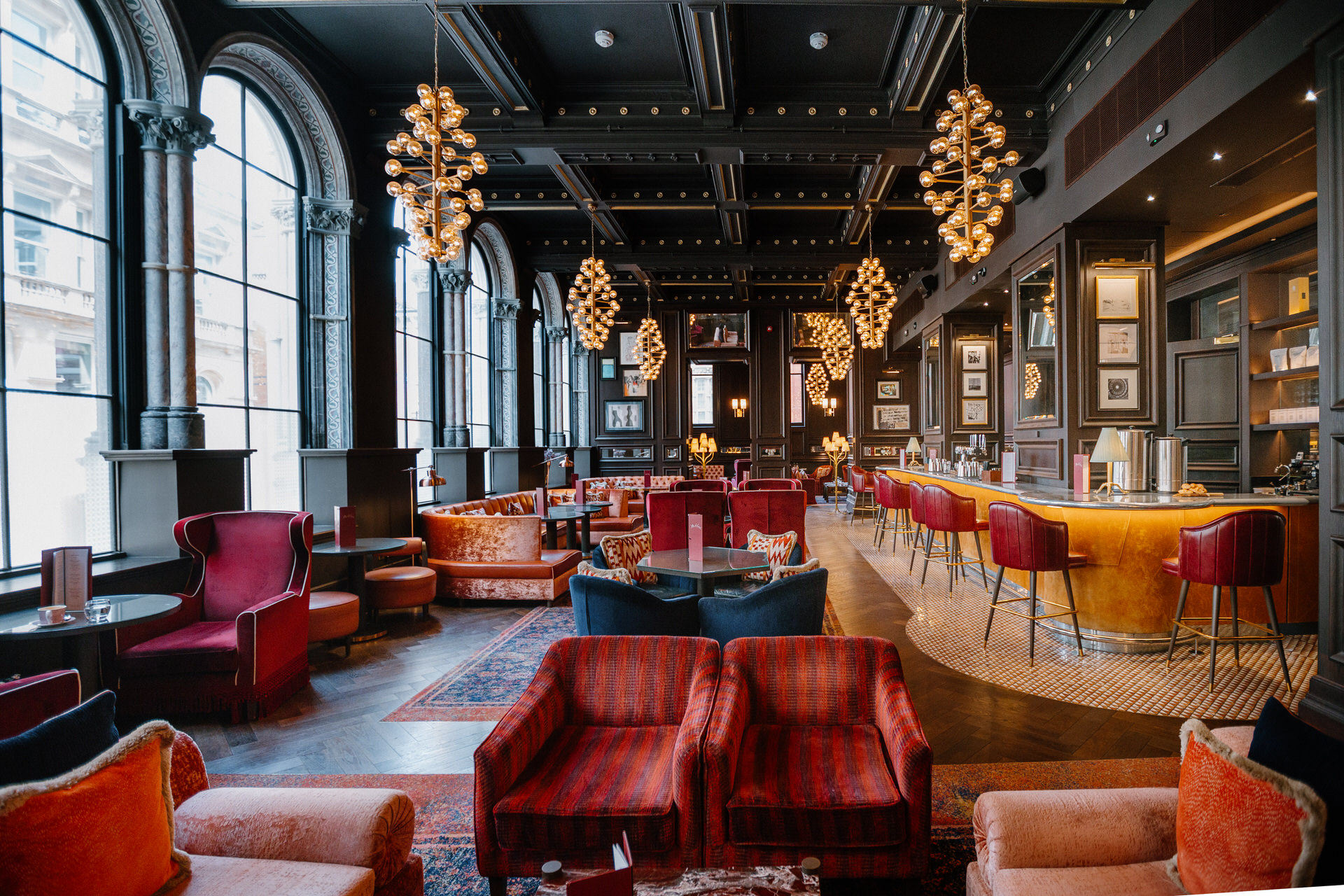 Elegant lounge with high arched windows, modern chandeliers, red and orange seating, and a long bar with gold accents in a stylish, upscale interior at The Grand Hotel Birmingham wedding venue