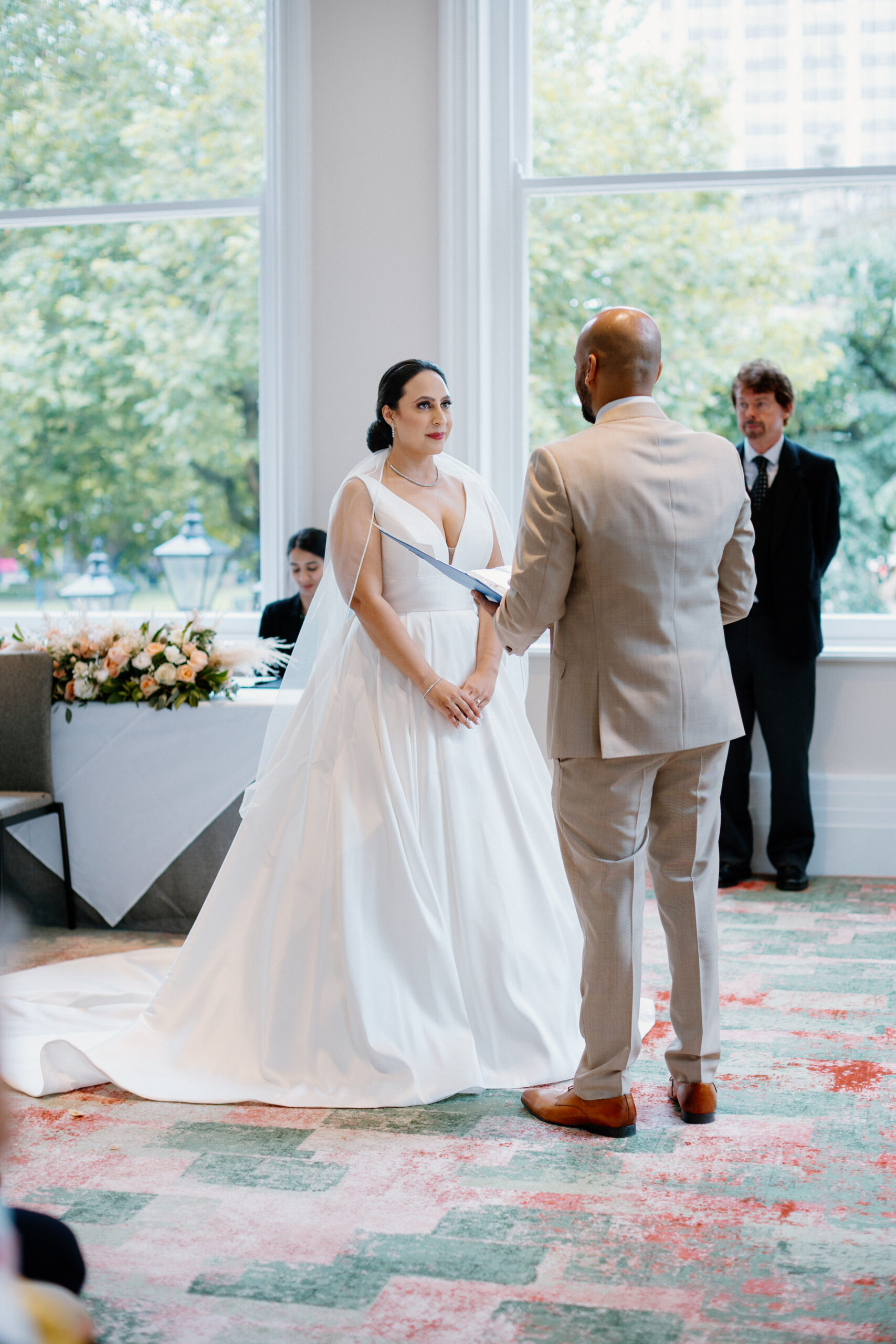 A bride and groom stand facing each other during their wedding ceremony in a bright room with large windows and floral decorations, beautifully captured by The Grand Hotel Birmingham Photographer.