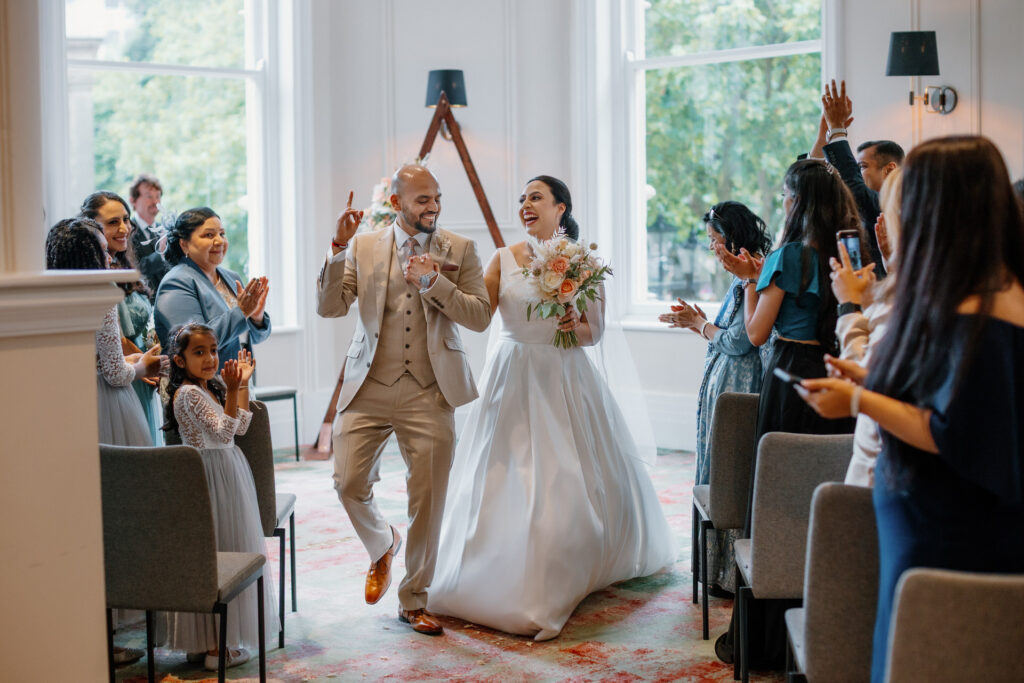 A newly married couple smiles and walks down the aisle, surrounded by cheering guests in a bright indoor setting.