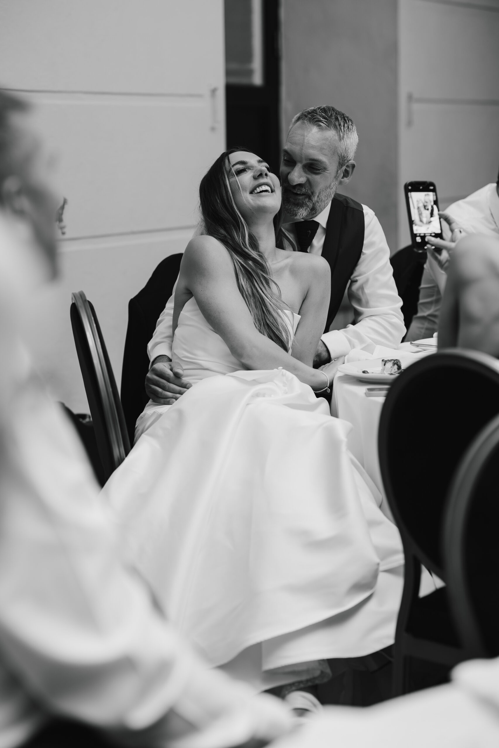 A bride in a strapless dress sits on a man's lap, both smiling, as a guest takes a photo with a phone at a wedding reception.