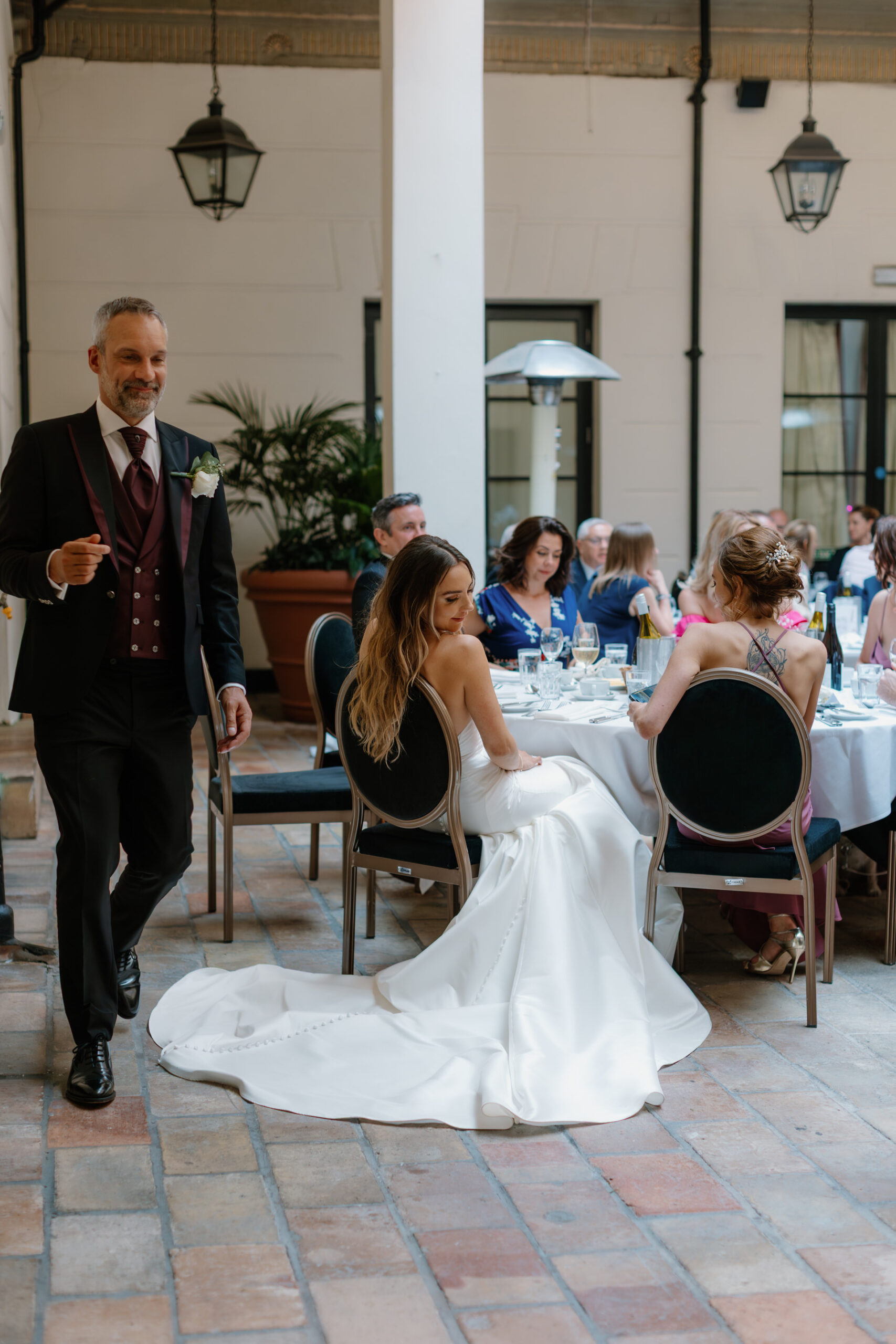 A bride in a white gown sits at a table during a formal reception, while a man in a suit walks by, and guests are seated around round tables.