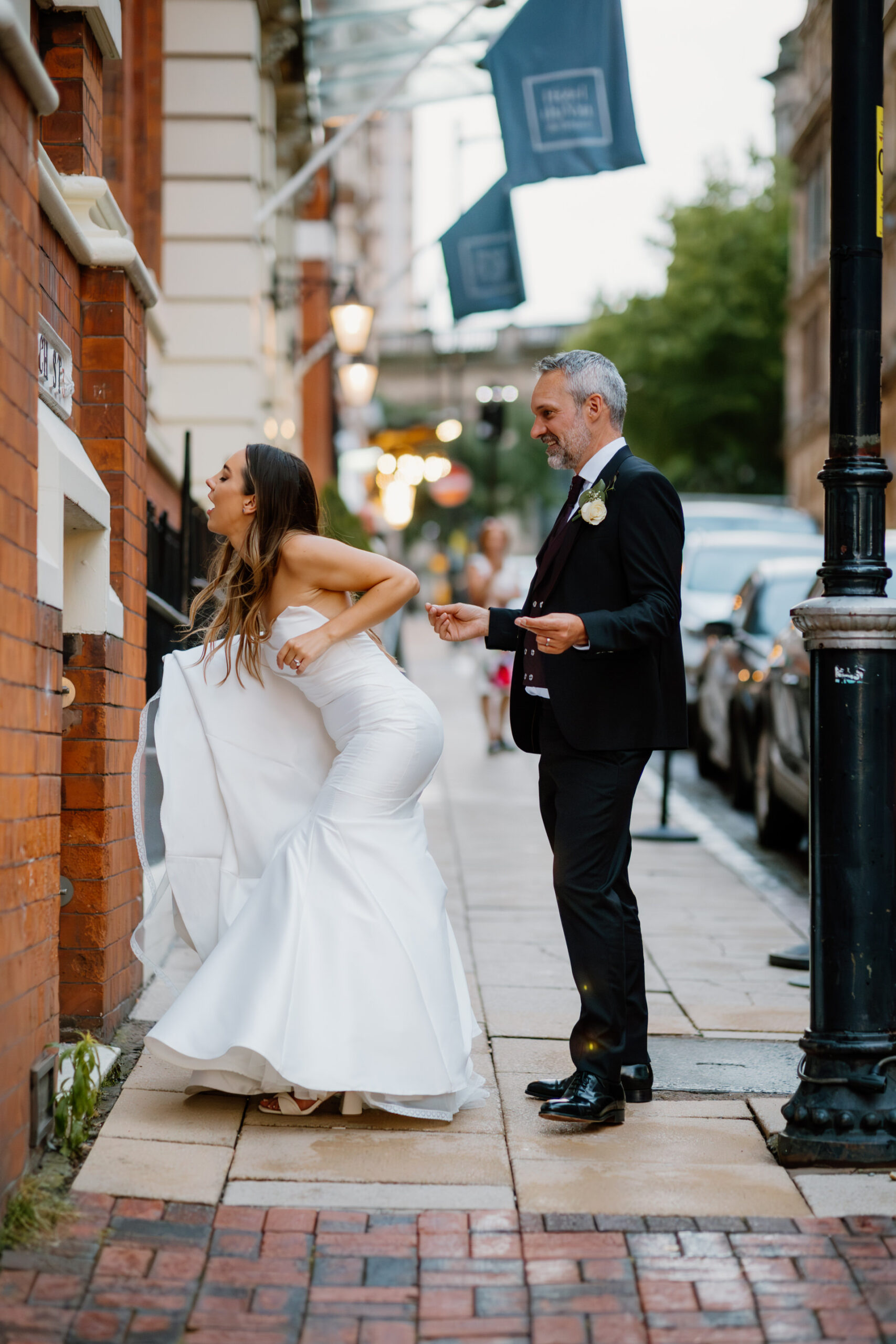 A bride in a white gown lifts her dress to adjust it on a city sidewalk while a man in a suit with a boutonniere stands nearby, holding a tablet and watching her.