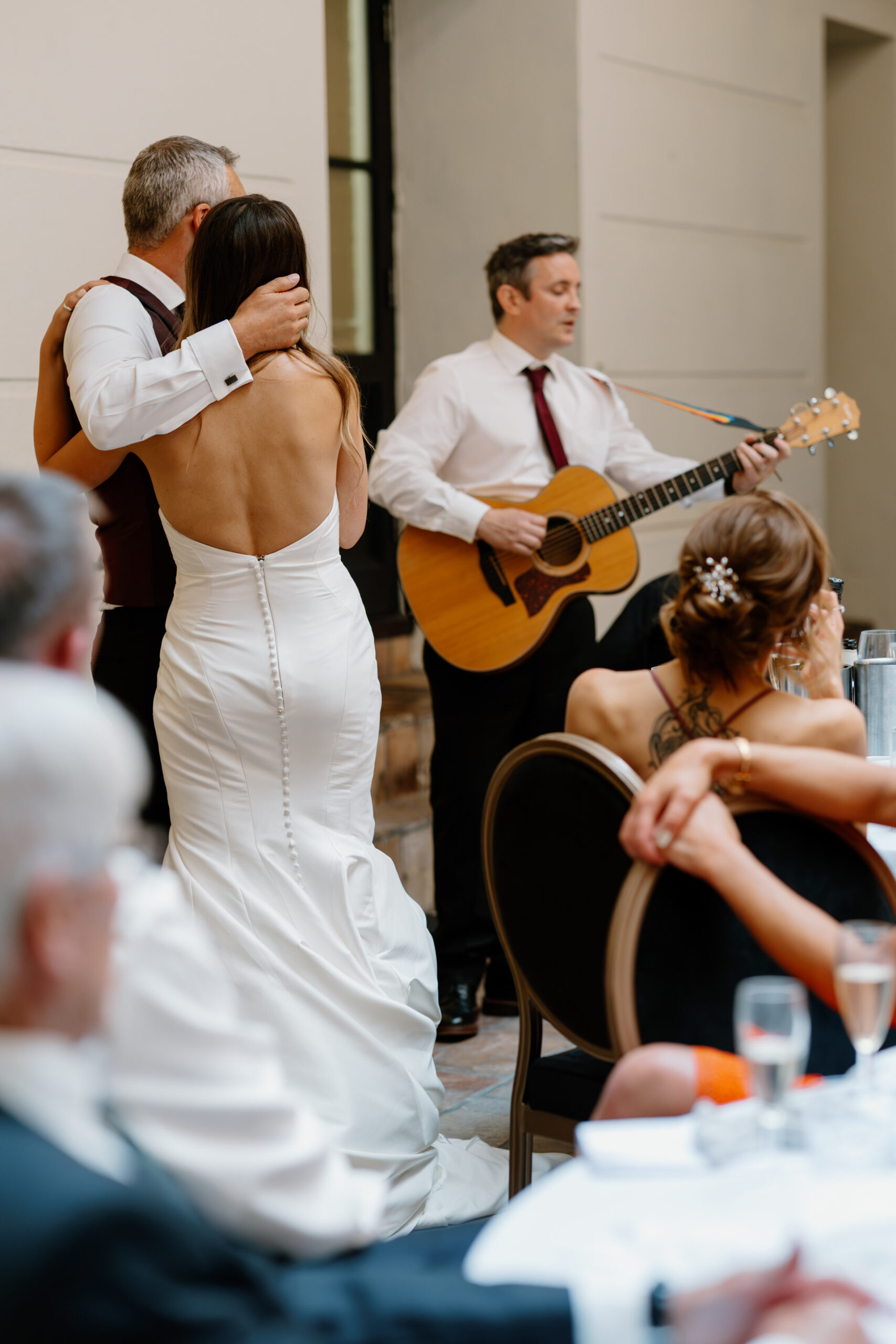 A bride in a white dress embraces an older man while a musician in a white shirt and tie plays guitar at a wedding reception. Seated guests watch the performance.