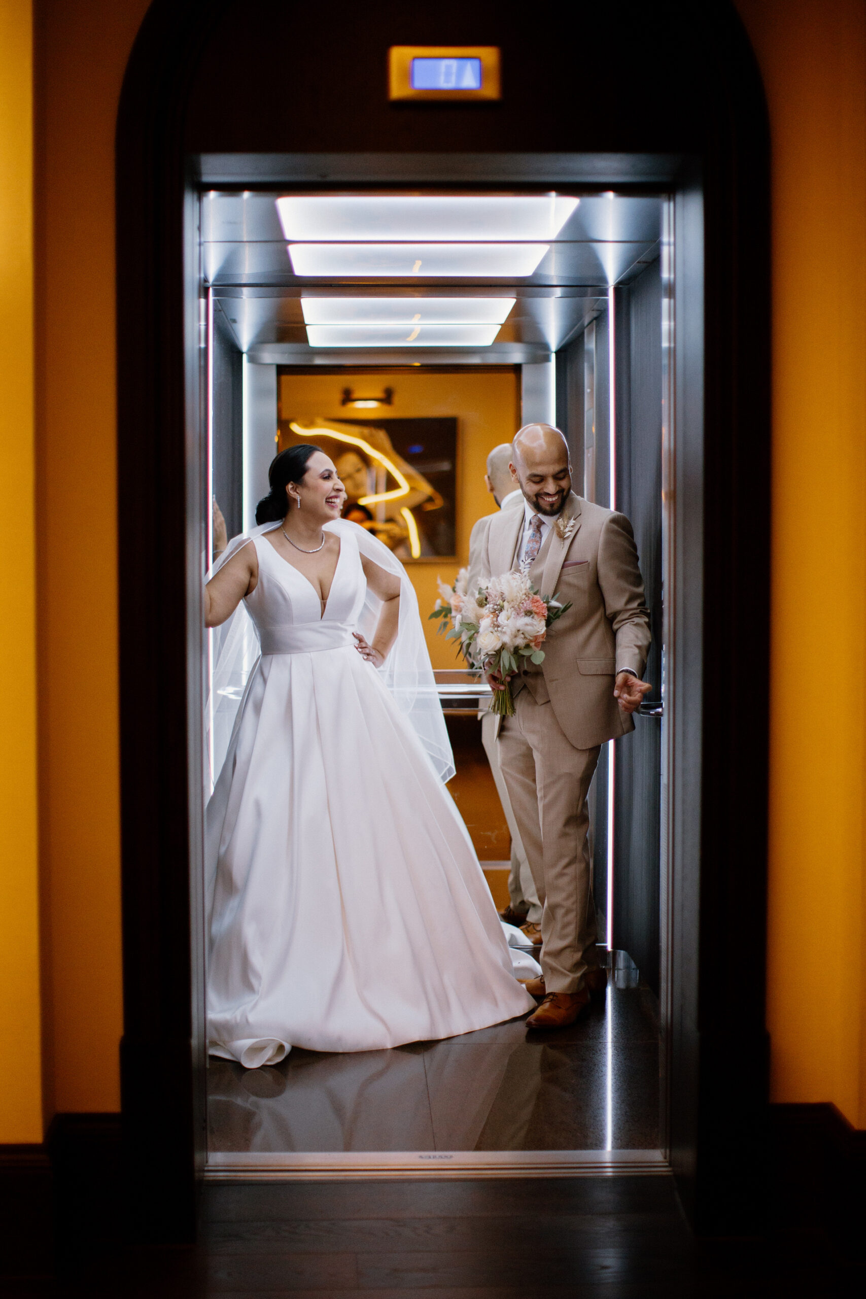 A bride in a white dress and a groom in a tan suit stand smiling inside a lit elevator, with the groom holding a bouquet of flowers.