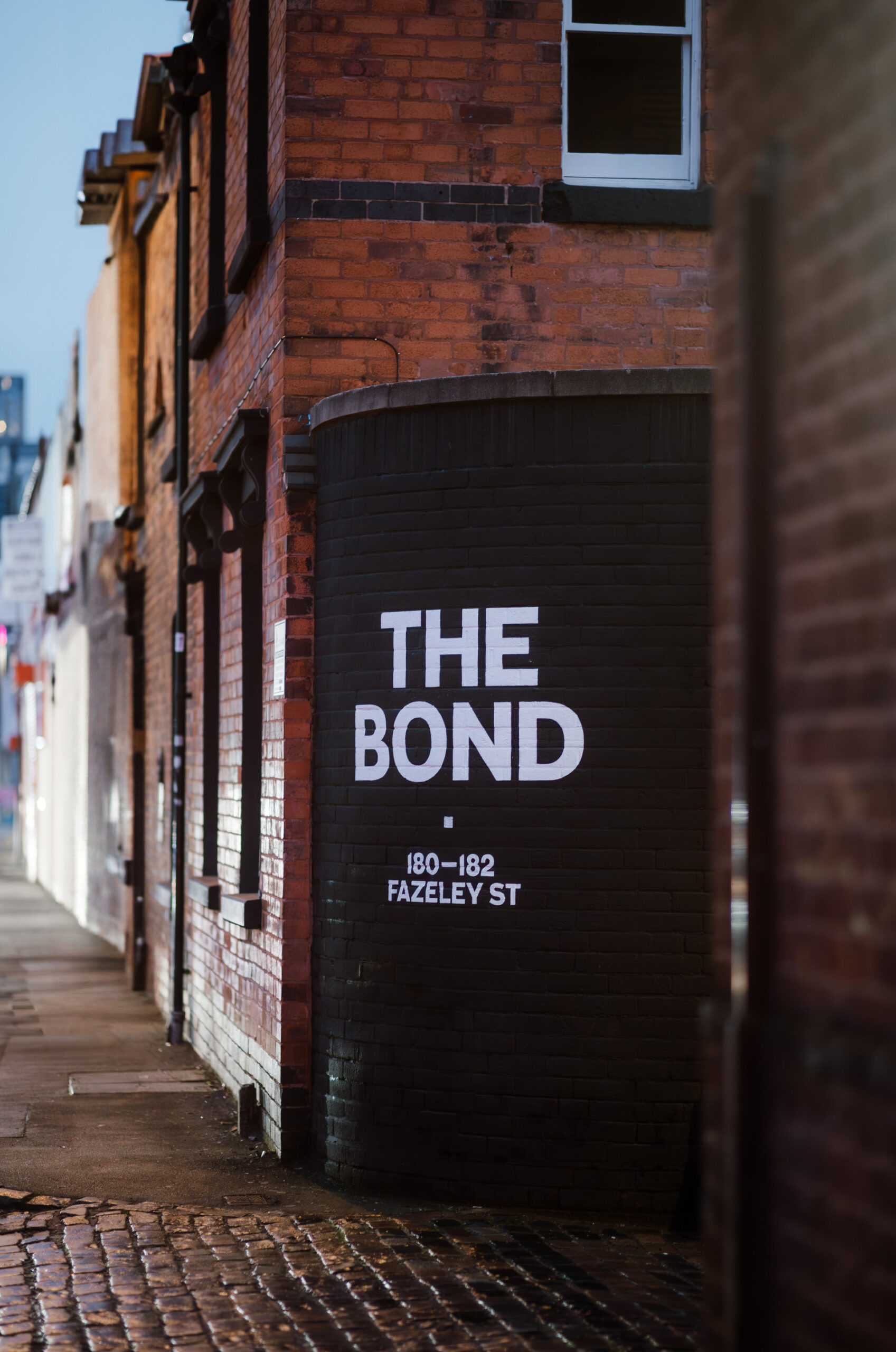 A brick building on a wet street corner displays the words "THE BOND 180-182 FAZELEY ST" painted in large white letters on a black wall.