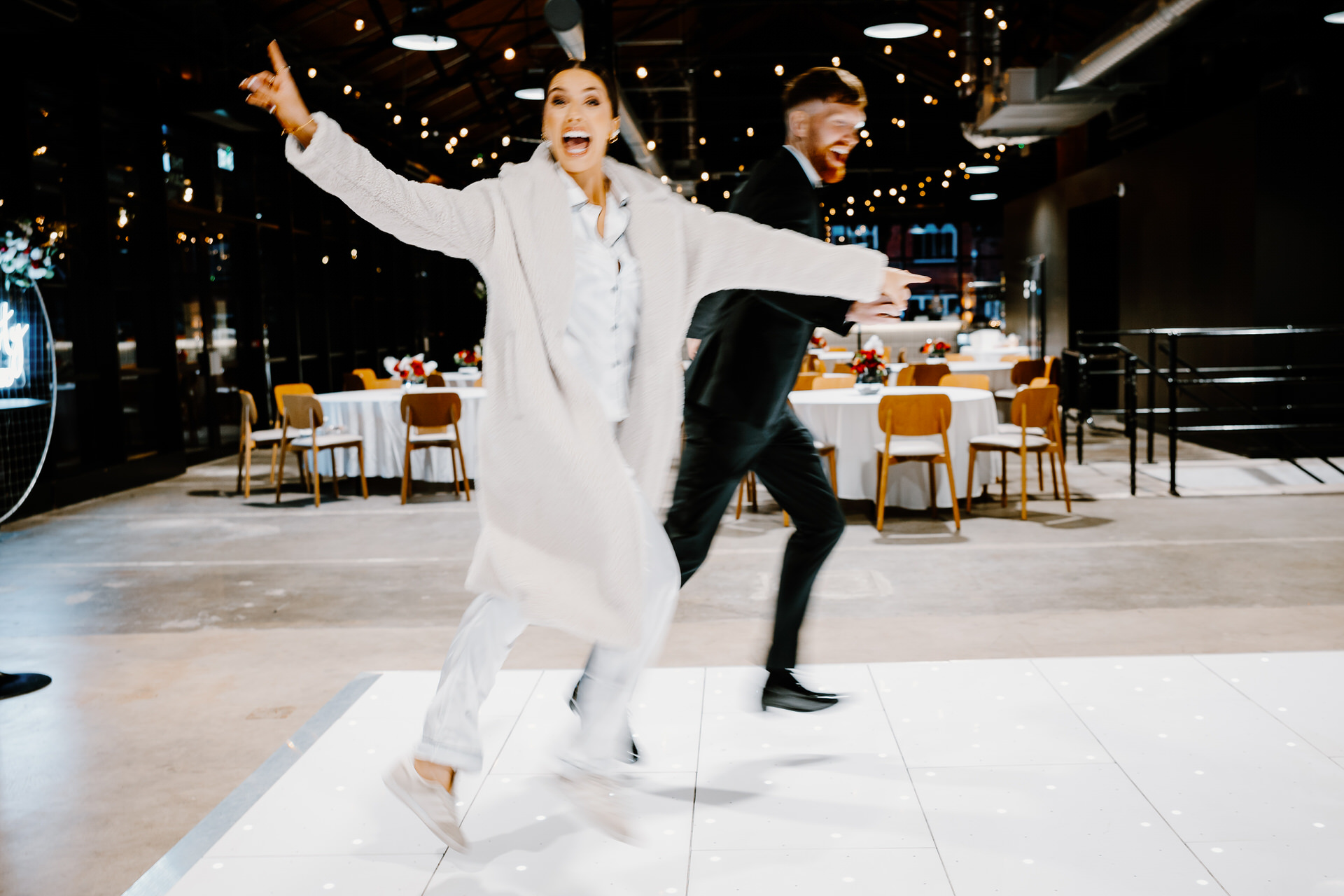 Woman in a light coat and man in a dark suit run playfully across a lit dance floor in a decorated indoor venue with tables and chairs in the background.