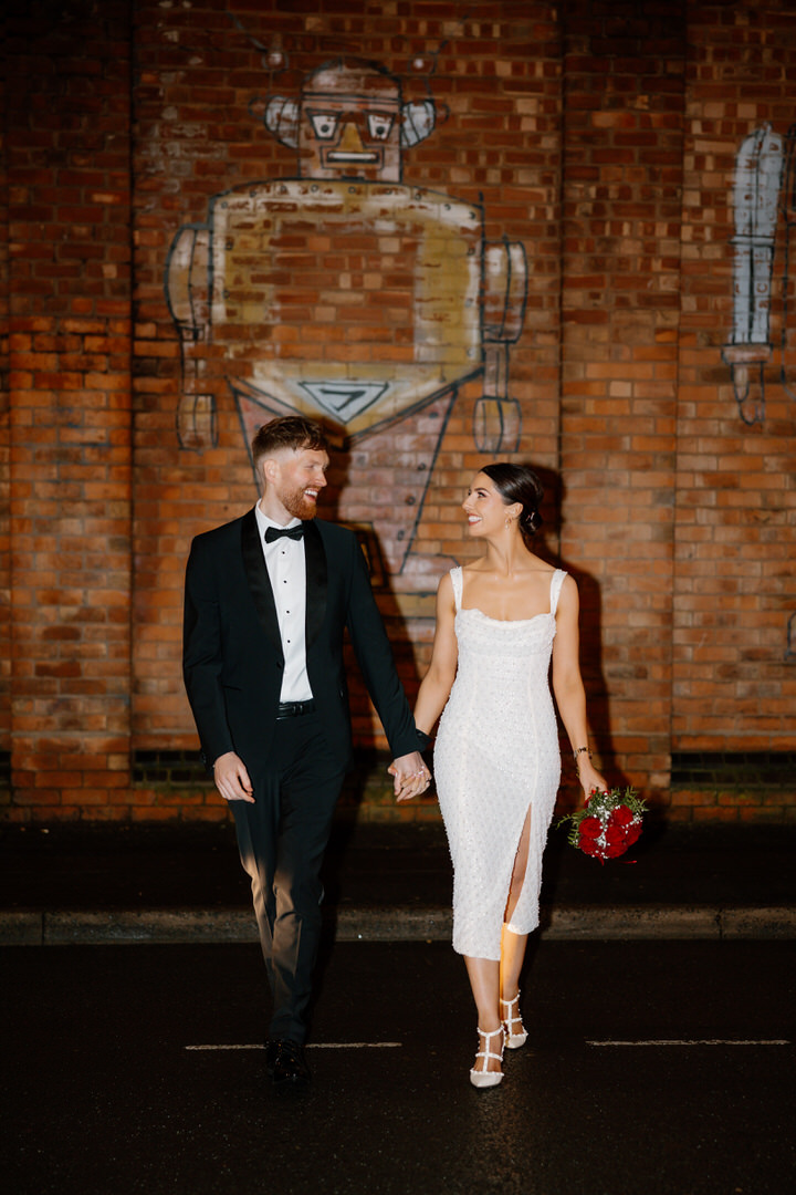 A couple dressed in formal attire walks hand in hand at night in front of a brick wall with a large robot mural. Captured by a Birmingham Digbeth wedding photographer, the woman holds a bouquet of red flowers.