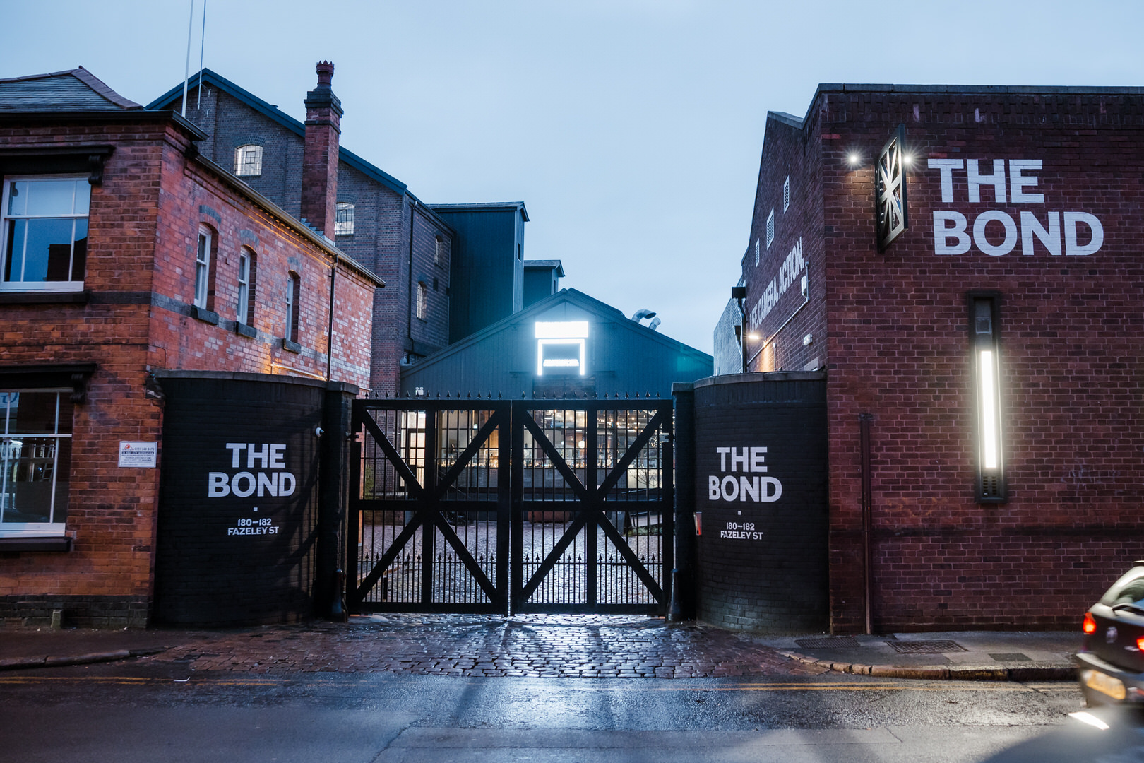 A gated entrance to a brick-building complex labeled "THE BOND" on both sides, seen at dusk with exterior lights on.
