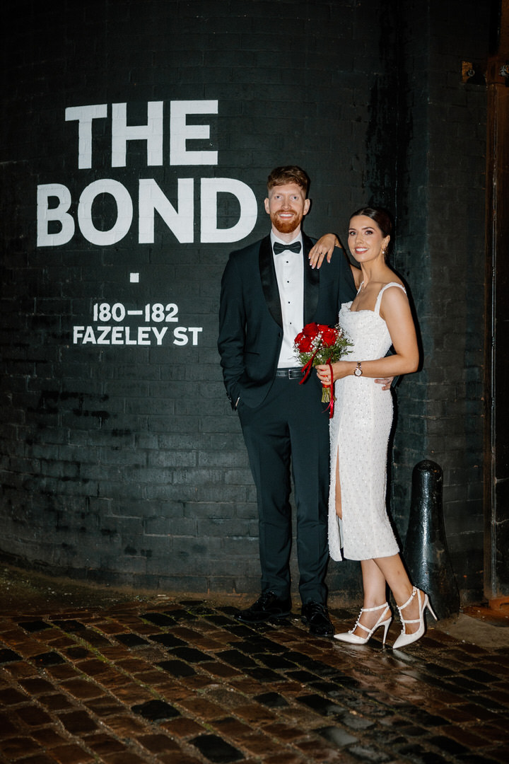 The bride and the groom pose and smile in front of a black wall with "THE BOND 180-182 FAZELEY ST" written in white letters, captured by a Digbeth Birmingham wedding photographer.