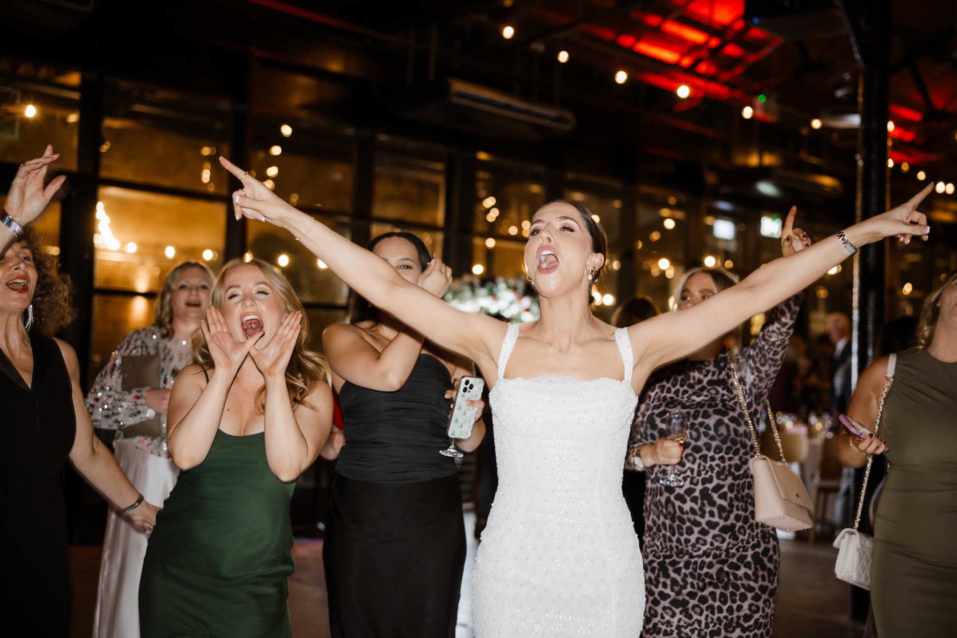 A bride in a white dress sings with arms raised, surrounded by cheering guests at an indoor wedding reception.