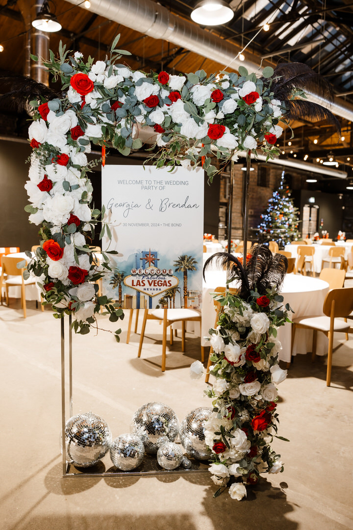 A floral wedding arch with red and white roses decorates a welcome sign for Georgina and Brendan’s wedding, with disco balls at the base and tables set up in the background.