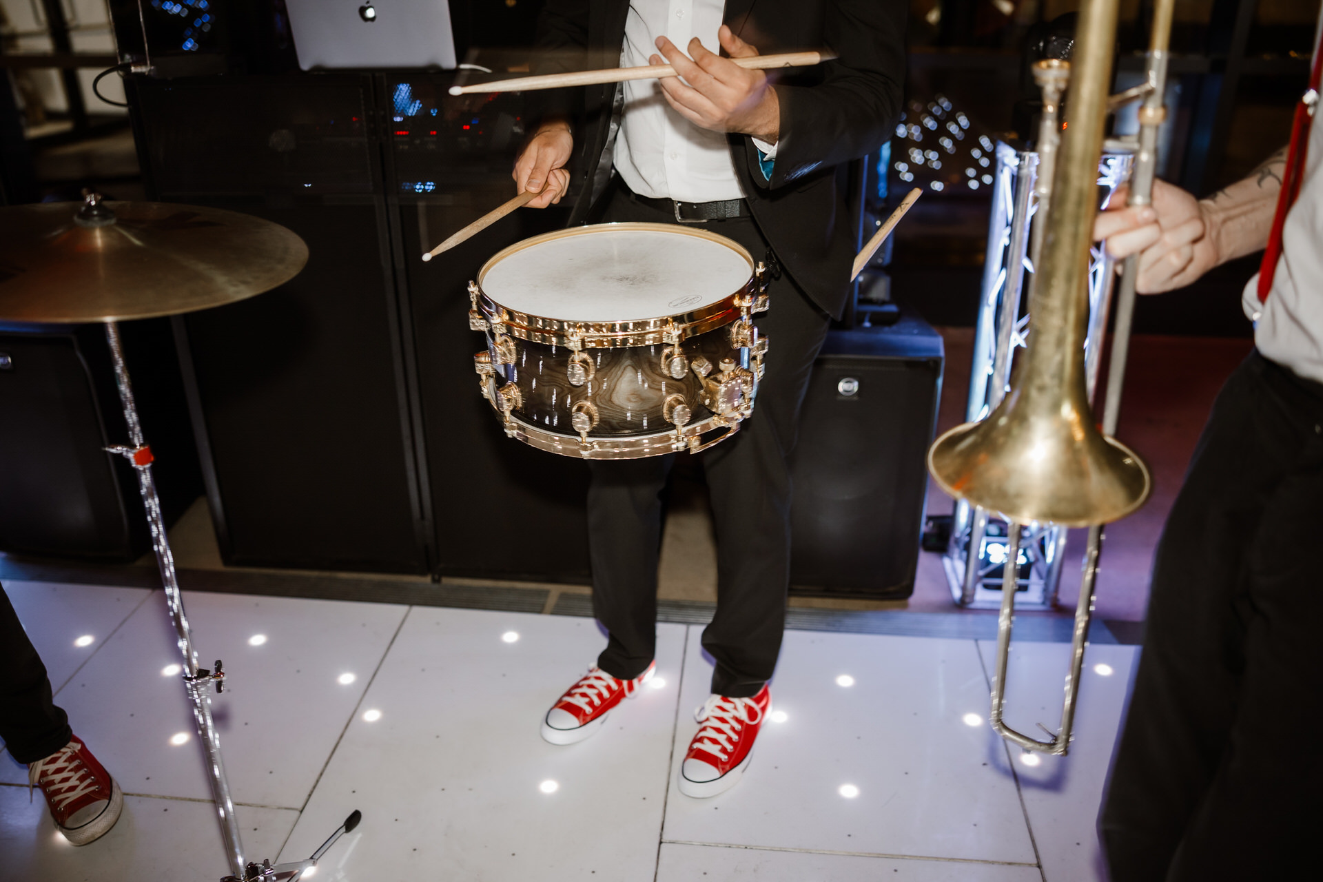 Person in a suit and red sneakers playing a snare drum, with nearby cymbal and trombone, on a white stage with lights.
