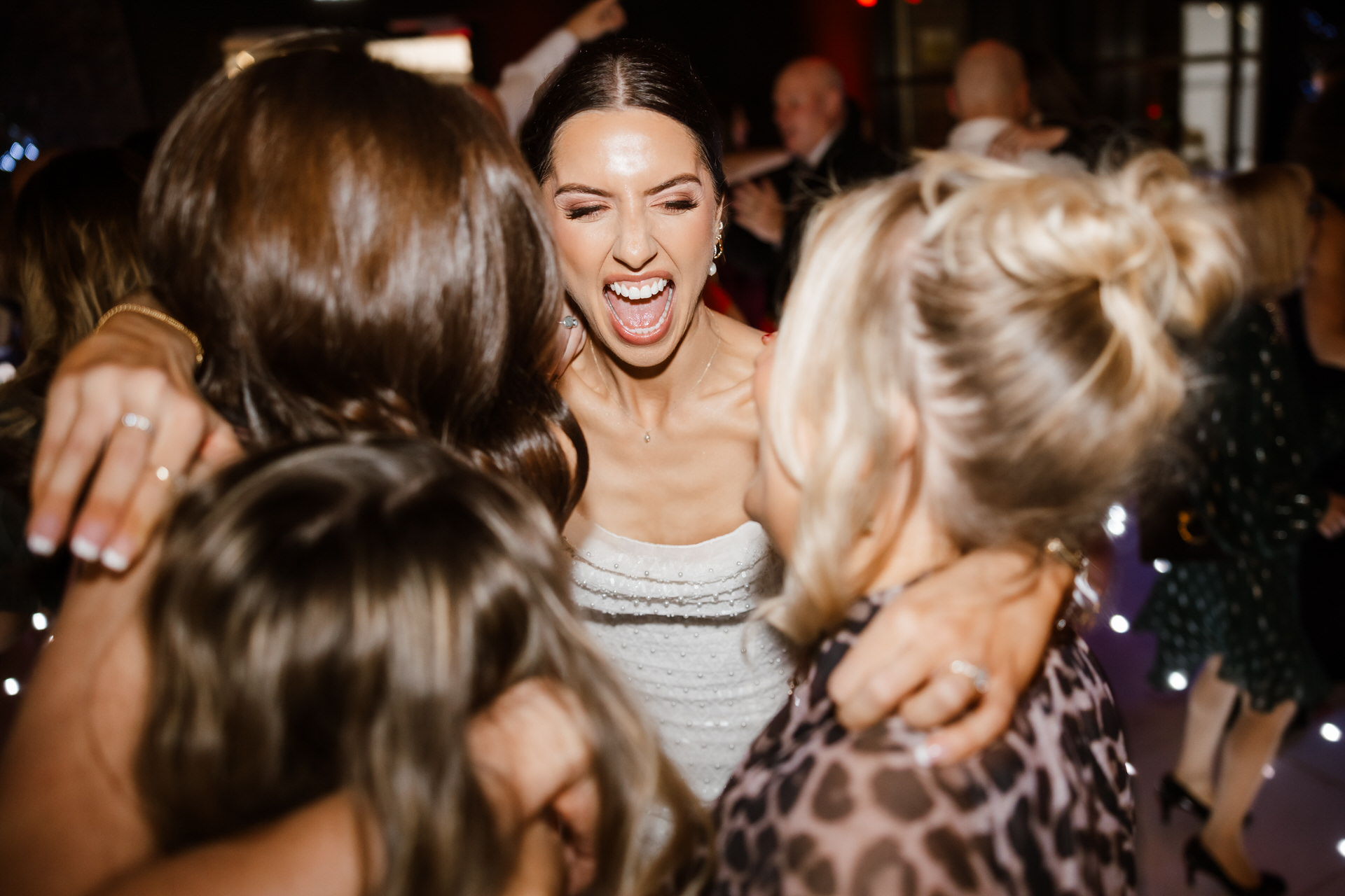 A woman in a white dress smiles and shouts while hugging three people in a crowded indoor setting.