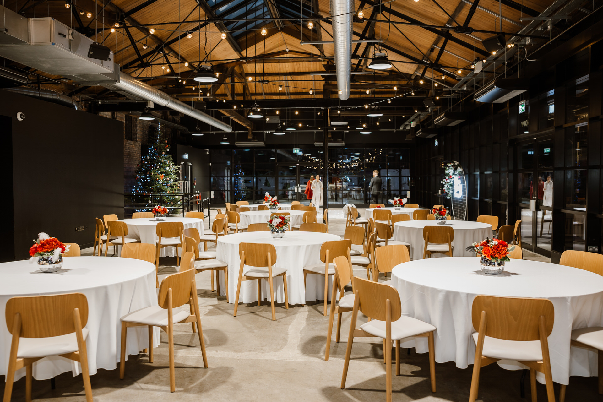 Spacious event hall at The Bond wedding venue in Birmingham, featuring round tables with white tablecloths, wooden chairs, floral centerpieces, twinkling string lights, and a decorated Christmas tree in the background.