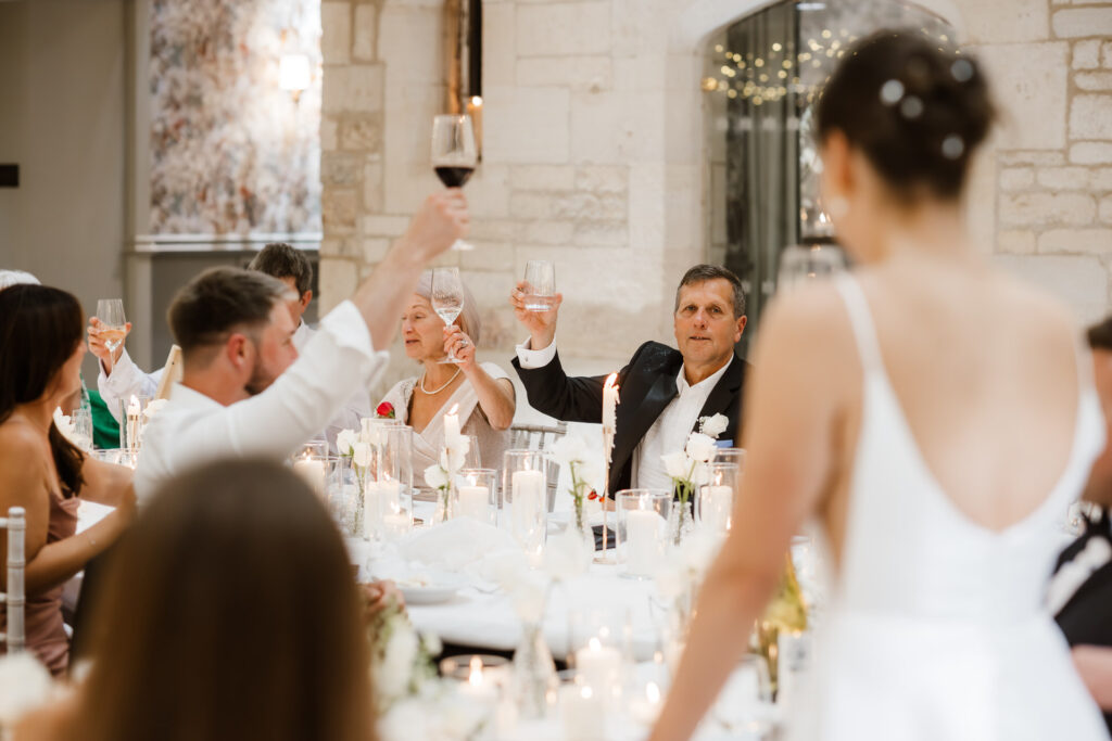 Guests at a formal event raise glasses in a toast around a decorated dining table, set in an elegant room with stone walls.