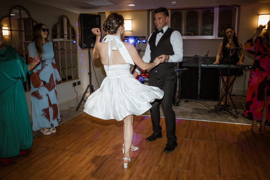 A couple in formal attire dances on a wooden floor, with the woman in a white dress spinning. Guests and a band are in the background.