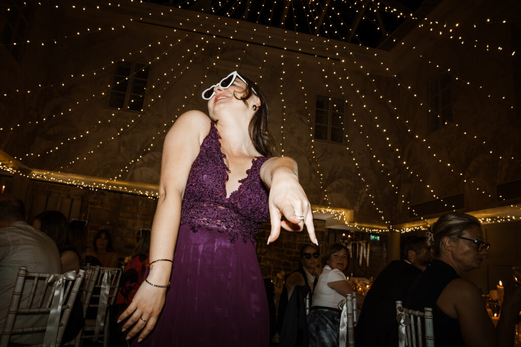 A woman in a purple dress dances enthusiastically at a decorated indoor event, pointing forward and wearing large white sunglasses.