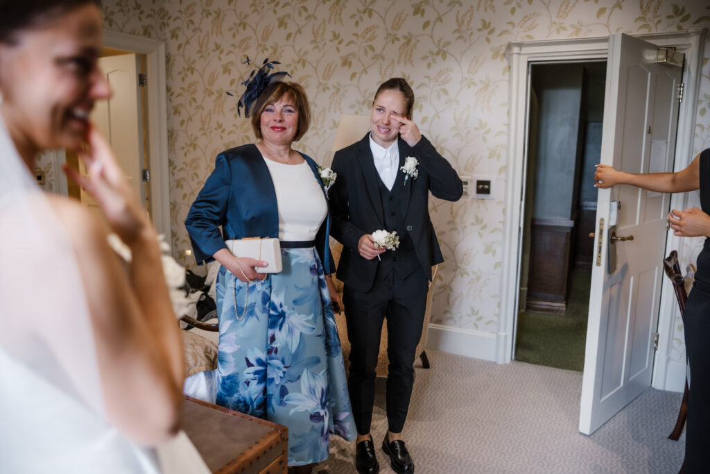 Two people smiling and holding flowers in a room with floral wallpaper. One is dressed in a blue jacket with a floral skirt, and the other in a dark suit. Another person is partially visible.