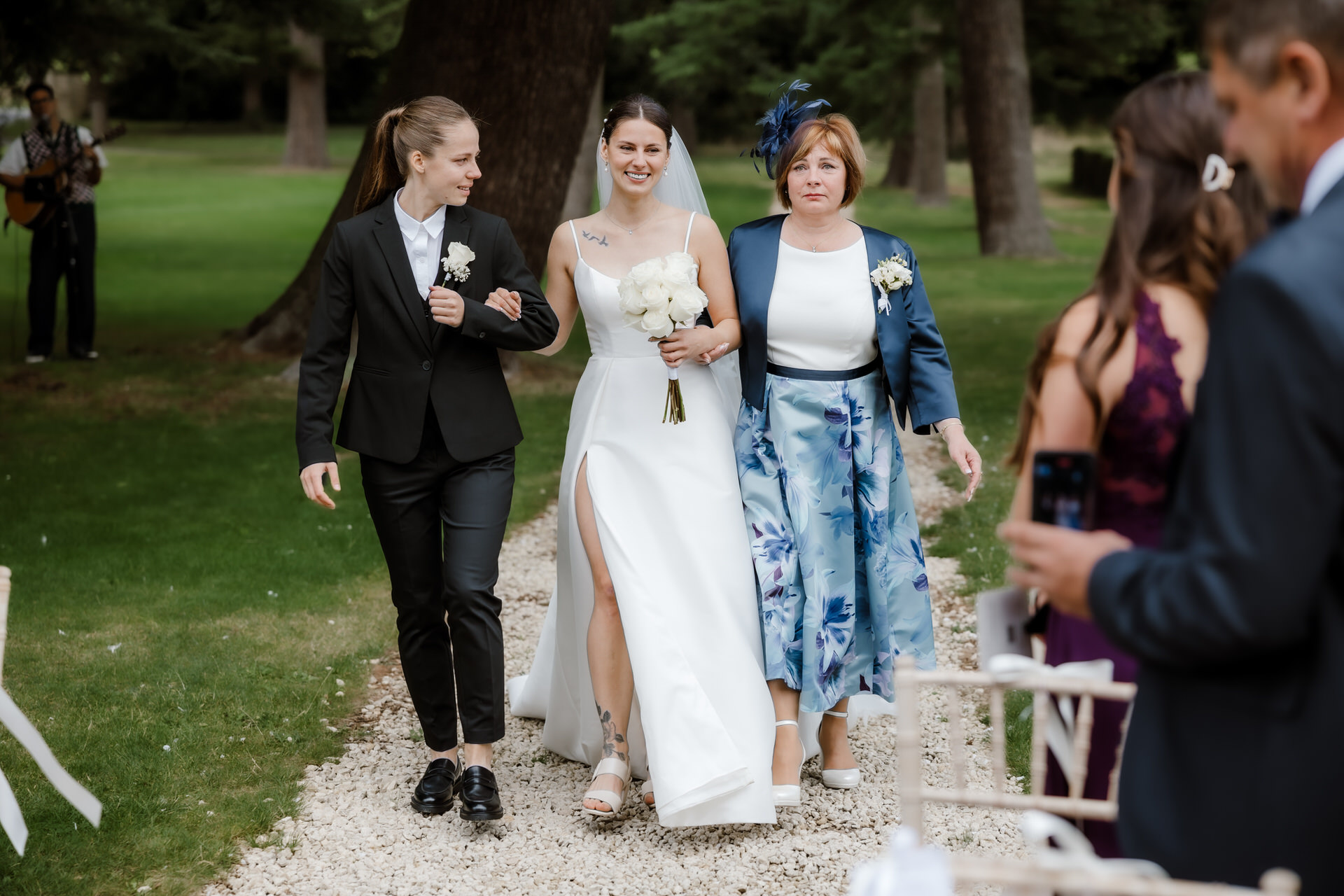 A bride in a white gown walks down the aisle outdoors, flanked by two women, one in a suit and the other in a floral dress. Guests are visible in the background.