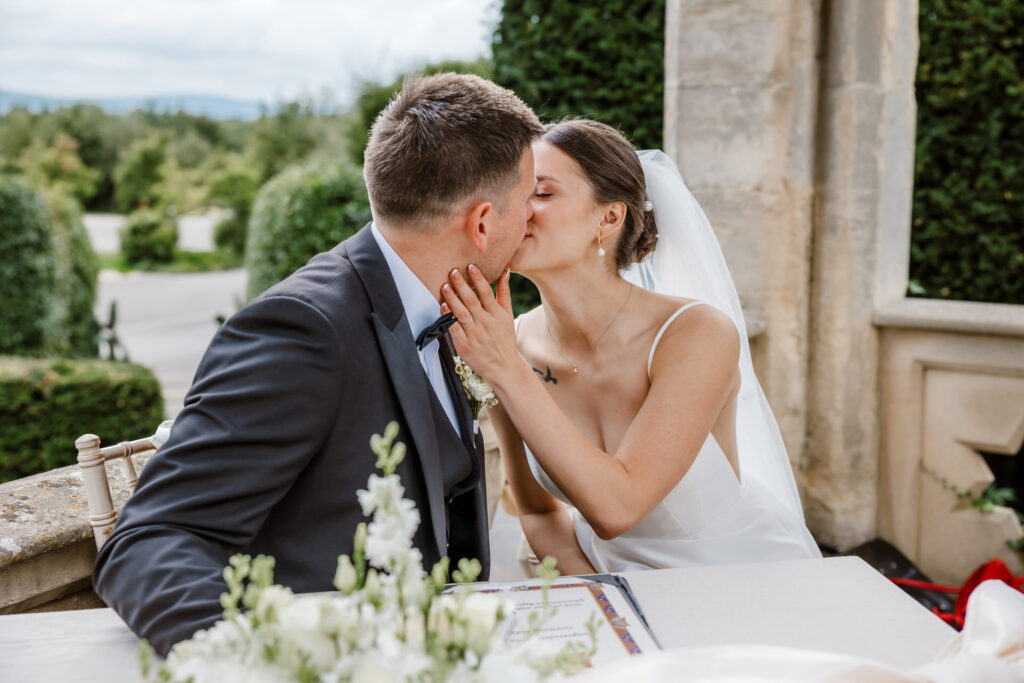 A bride and groom share a kiss while seated at a table outdoors, with flowers and greenery in the background.