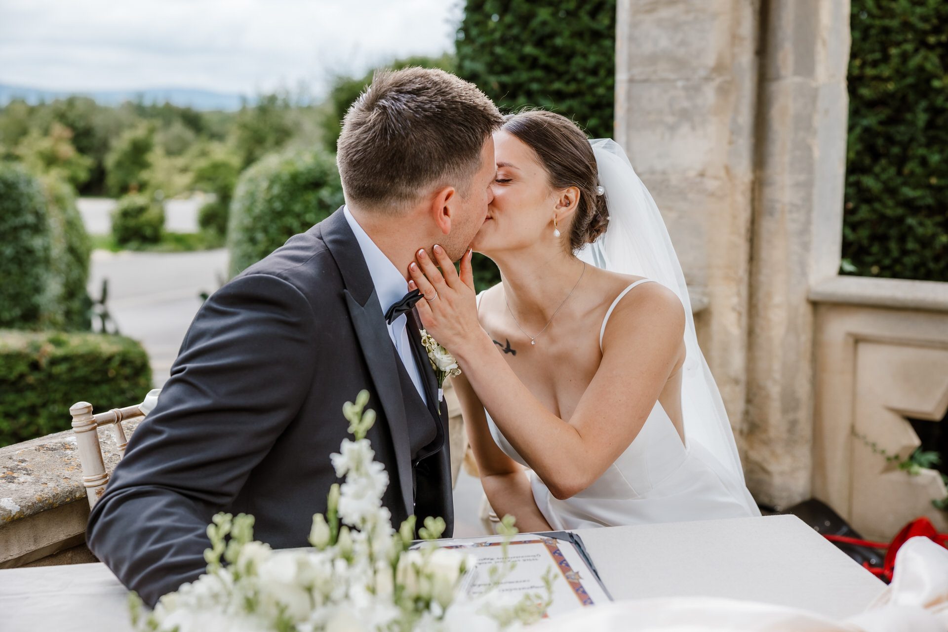 Ellenborough Park Wedding Photographer 51 A bride and groom share a kiss while seated at a table outdoors, with flowers and greenery in the background.