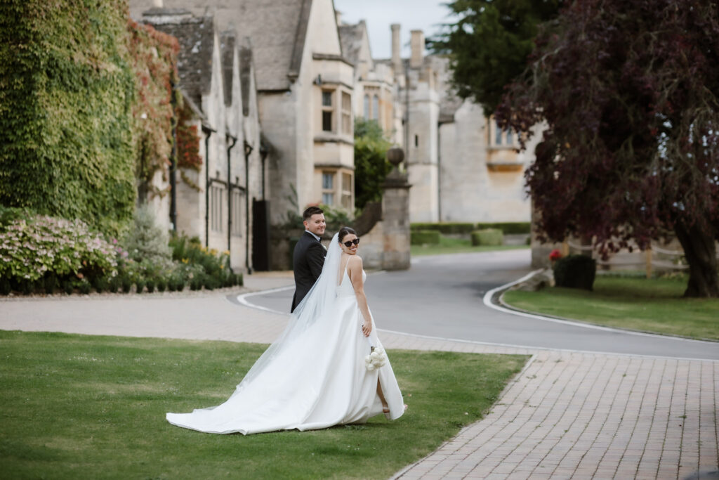A bride and groom walk along a path by the historic, ivy-clad stone building of Ellenborough Park. She wears a white gown and veil, holding a bouquet under the overcast sky, creating a perfect scene for wedding photography.