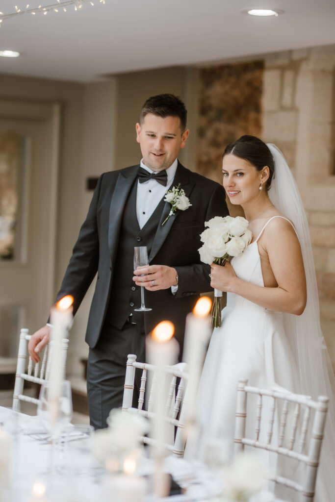 A bride in a white gown holds a bouquet, standing with a groom in a tuxedo, near a candlelit table in an elegant setting.