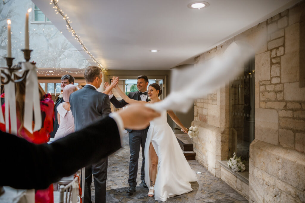 Bride and groom joyfully walking hand in hand through a hallway lined with guests, some waving handkerchiefs.